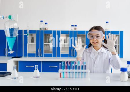 ragazza sorridente in occhiali con le mani in guanti in lattice vicino a matracci e provette in laboratorio, immagine stock Foto Stock