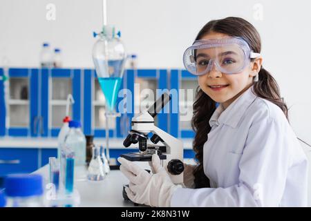 presen ragazza in occhiali e cappotto bianco sorridente alla macchina fotografica vicino al microscopio in laboratorio, immagine di scorta Foto Stock