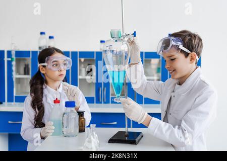 ragazzo sorridente che tiene fiasca con liquido vicino a ragazza seria in occhiali, immagine di scorta Foto Stock