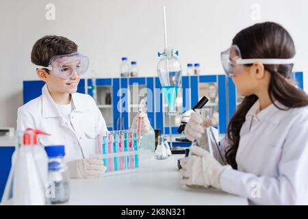 ragazzo sorridente in occhiali guardando la ragazza offuscata vicino a provette e matracci in laboratorio, immagine stock Foto Stock