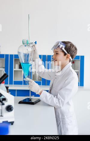 vista laterale di un bambino sorridente in guanti di lattice che guarda una fiasca con liquido blu vicino al microscopio, immagine stock Foto Stock