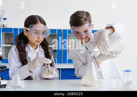 tensed ragazza in occhiali guardando il ragazzo sorridente versando liquido in fiasca mentre facendo esperimento chimico, immagine di scorta Foto Stock