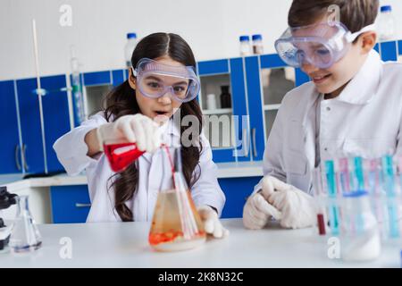 ragazza stupita in occhiali che versano liquido in fiasca durante esperimento chimico vicino amico sorridente, immagine di scorta Foto Stock