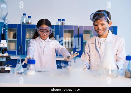 ragazza in occhiali in piedi con le mani estese vicino fiasche di vapore e sorridente amico con il viso sporco, immagine stock Foto Stock