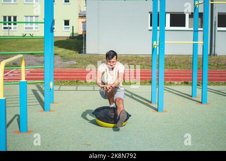 Un atleta professionista dai capelli castani fa una squadra a una gamba su un fascio di bilanciamento per migliorare le capacità motorie. Parco giochi all'aperto. Prepararsi per il mare Foto Stock