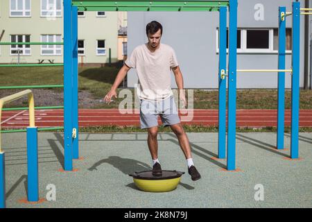 Un atleta professionista dai capelli castani fa una squadra a una gamba su un fascio di bilanciamento per migliorare le capacità motorie. Parco giochi all'aperto. Prepararsi per il mare Foto Stock