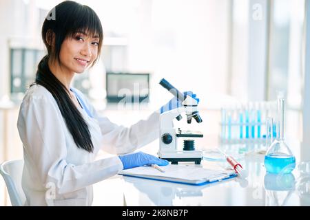 Sorridente scienziata asiatica in posa in moderno laboratorio di ricerca medica accanto al microscopio. Foto Stock