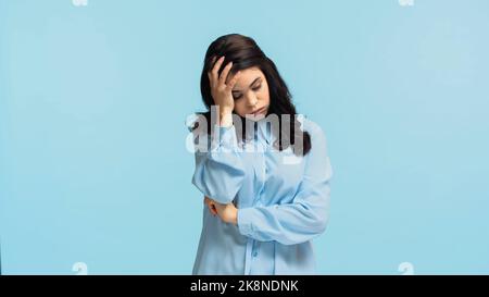 giovane donna dispiacente in camicia che tocca la testa e puffing guance isolato su blu Foto Stock