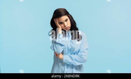 giovane donna dispiacente in camicia che tocca la testa mentre puffing guance isolato su blu Foto Stock