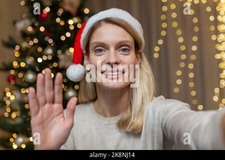 Donna matura a casa per festeggiare il Capodanno e il Natale, bionda che parla in videochiamata utilizzando l'app sul telefono per comunicare con gli amici in remoto, moglie vicino all'albero di Natale nel soggiorno di casa. Foto Stock