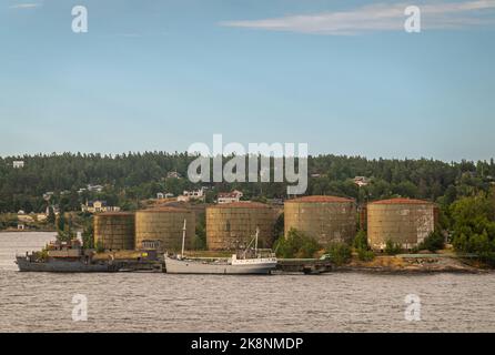 Svezia, Stoccolma - 16 luglio 2022: Primo piano, vecchio porto petrolifero sull'isola di Stora Hoggarn mostra serbatoi cilindrici marroni e un molo con 2 navi più vecchie sotto Foto Stock