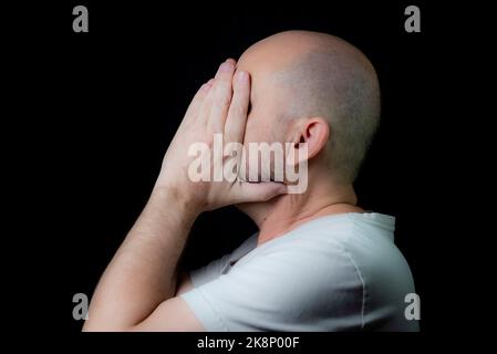 Un ritratto di un calvo frustrato uomo caucasico con le mani sul volto contro lo sfondo nero Foto Stock