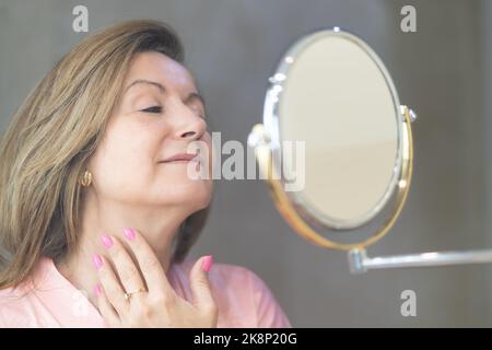 Donna anziana che si prende cura della sua pelle di fronte a uno specchio in bagno. Foto Stock