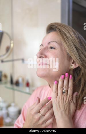 Donna bianca adulta che applica cosmetici e crema sul viso e sul collo per la cura della pelle. Foto Stock