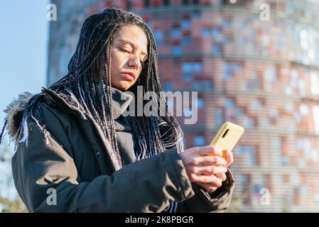 Donna ispanica focalizzata in outerwear con dreadlocks messaggi di testo sul cellulare moderno, mentre in piedi sulla strada vicino a un edificio in città Foto Stock