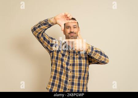 Bel maschio felice bearded gli occhi e guarda attraverso una cornice formata dalle sue mani. Un uomo attraente fa una cornice con le sue dita, isolato Foto Stock