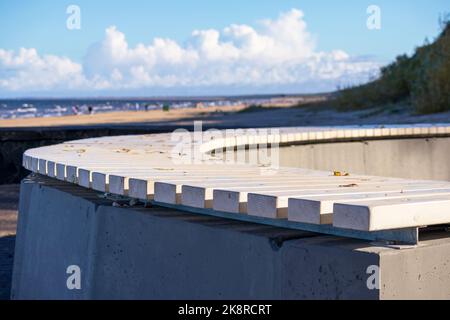 Una panca semicircolare di legno bianco sulla spiaggia di sabbia con cielo blu e alcuni alberi verdi nel campo del becakground Foto Stock