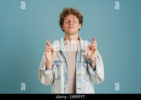 Giovane uomo riccio che indossa una giacca sorridente e che tiene le dita incrociate isolate su sfondo blu Foto Stock