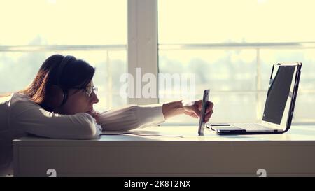 Giovane donna pigra, lavoratore in ufficio che indossa le cuffie, gioca ai giochi per dispositivi mobili con lo smartphone durante l'orario di lavoro. Dipendente annoiato. Lavoratore inutile e improduttivo non facendo nulla e dimenticare il suo lavoro. Foto di alta qualità Foto Stock