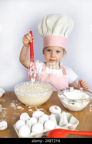 Una ragazza nel cappello di uno chef prepara la pasta, nelle sue mani una frusta Foto Stock