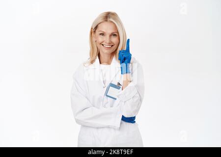 Bel medico sorridente, dermatologo donna in guanti medici, mostra un dito, prima regola, in piedi su sfondo bianco Foto Stock