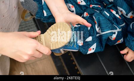 La madre mette delle scarpe blu sul bambino bambino del bambino del piede che siede nel corridoio domestico. Donna mamma vestirsi caldo stivali vestiti su bambino per la passeggiata invernale in c Foto Stock