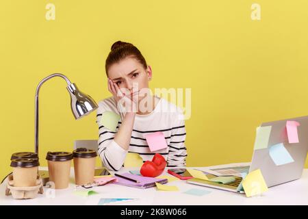 Annoiato donna impiegato ufficio coperto con appiccicoso seduta sul posto di lavoro appoggiando testa sulle mani, solo deluso donna stanca di lavoro wimmp. Note Studio interno girato isolato su sfondo giallo Foto Stock