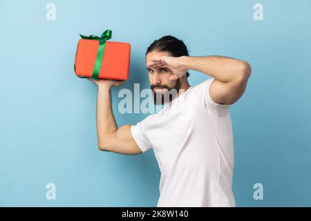 Ritratto dell'uomo con barba che indossa una T-shirt bianca con scatola regalo, guarda lontano con la mano sopra gli occhi, attende le vacanze future, festeggiamenti di compleanno. Studio in interni isolato su sfondo blu. Foto Stock