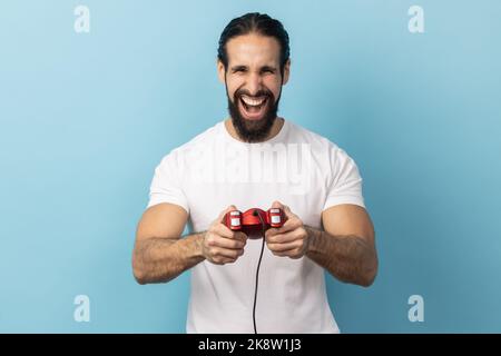 Ritratto dell'uomo con barba che indossa una T-shirt bianca che tiene in mano il joystick rosso gamepad, guardando la fotocamera con espressione ottimista, godendo di gioco. Studio in interni isolato su sfondo blu. Foto Stock