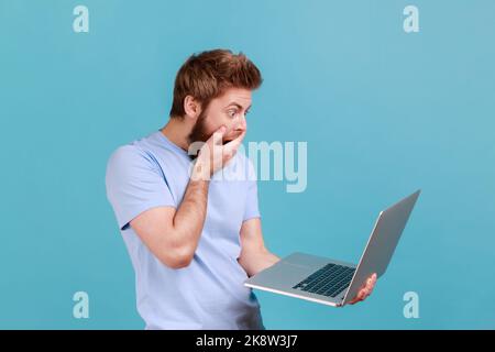 Ritratto di bell'uomo sorpreso bearded guardando il display del notebook con grandi occhi scioccati di contenuti, bocca coperta di palmo. Studio in interni isolato su sfondo blu. Foto Stock