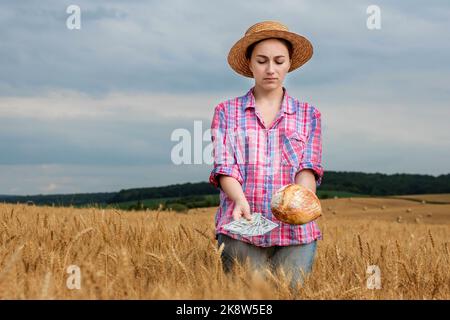 Giovane agricoltore che tiene dollari e pane in campo di grano dorato. Profitto dall'agricoltura durante la stagione di raccolta in estate. Foto Stock
