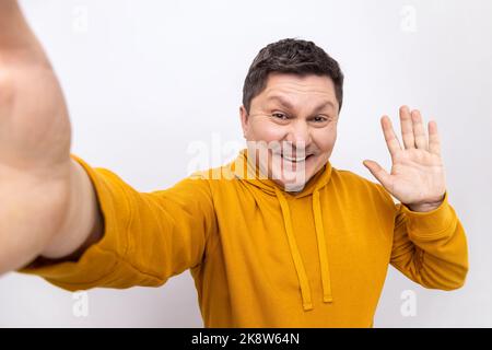 Ritratto di un bell'uomo positivo che fa selfie o streaming, mano ondulata POV, punto di vista della foto, indossando felpa con cappuccio stile urbano. Studio in interni isolato su sfondo bianco. Foto Stock