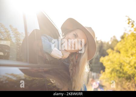 Sognando donna carina con gioia lasciare la città che si sta attaccando testa fuori dal finestrino dell'auto aperto. Giovane donna bionda in cappello con umore felice andare a viaggio in auto. In viaggio Foto Stock