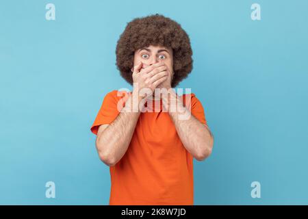 Uomo con acconciatura Afro in T-shirt arancione che chiude la bocca per non urlare, sentendosi spaventato e terrorizzato, i suoi occhi e guardare pieni di paura e terrore. Studio in interni isolato su sfondo blu. Foto Stock