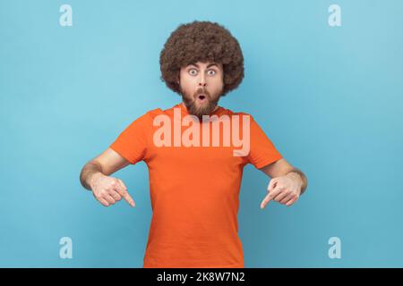 Ritratto dell'uomo sorpreso scioccato con acconciatura afro che indossa una T-shirt arancione rivolta verso il basso con le dita anteriori, guardando la fotocamera a bocca aperta. Studio in interni isolato su sfondo blu. Foto Stock