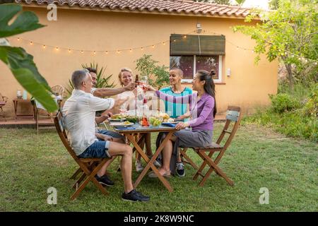 Amici multietnici che brinda sorridendo e ridendo felici nel patio. Gente allegra di mezza età intorno al tavolo. Foto Stock