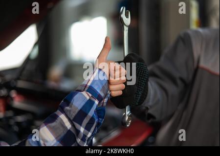 Primo piano della mano della donna pollice su e mano del meccanico con l'attrezzo. Il master automobilistico nei guanti mostra un gesto positivo dopo aver terminato il lavoro Foto Stock