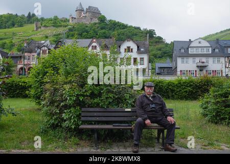 Il vecchio con il cappuccio piatto, i baffi del manubrio e il binocolo siede sulla panchina nella pittoresca Bacharach, Germania. Viaggi anziani agili sani in pensione. Foto Stock