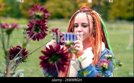 Donna che scatta foto con lo smartphone nel parco. Positivo giovane ragazza hipster con lunghi dreadlocks in abiti casual fotografando fiori su cellulare phon Foto Stock
