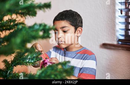 Allestire per Natale. Un ragazzino spensierato che mette le decorazioni natalizie su un albero a casa durante il periodo natalizio. Foto Stock