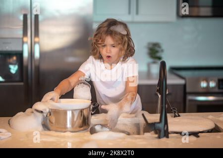 Divertenti ragazzi gemelli che aiutano in cucina con piatti di lavaggio. Bambini che si divertono con i lavori di casa. La cameriera dei bambini lava i piatti con acqua saponata. Carino Foto Stock