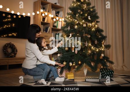 Felice piccola prescolare ragazza caucasica bambino decorazione albero di Natale con felice madre giovane, mettendo giocattoli sui rami, godendo di preparare per la celebrazione di Capodanno a casa, il concetto di tempo di miracolo. Foto Stock