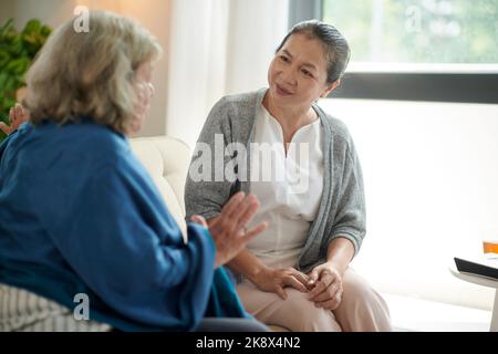 Donna anziana eccitata che parla con il migliore amico mentre si siede sul divano nel soggiorno Foto Stock