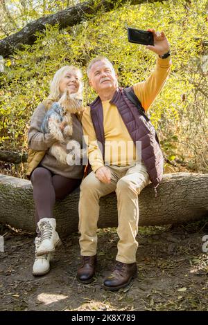 Foto verticale di una coppia matura e di un cane dello Yorkshire che prende selfie mentre si siede nella foresta Foto Stock