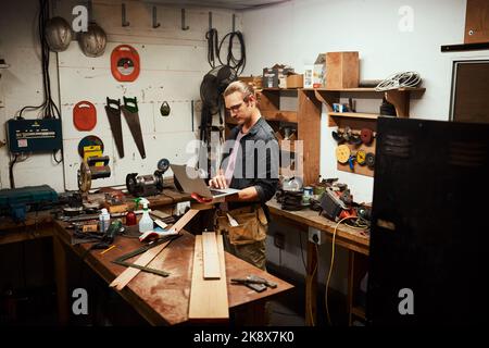 Alla ricerca di ispirazione. Un giovane falegname maschio concentrato che lavora sul suo computer portatile mentre si trova all'interno del suo laboratorio di notte. Foto Stock