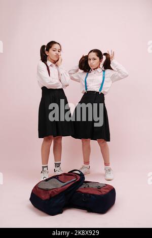 Due studentesse gemelle con guance a coda doppia che si spugnano per una foto. Gli zaini sono impilati in primo piano. Indossare uniformi scolastiche con skir alto al ginocchio Foto Stock