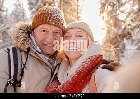 Ritratto POV di felice coppia anziana scattando foto selfie mentre si gode l'escursione nella foresta invernale Foto Stock