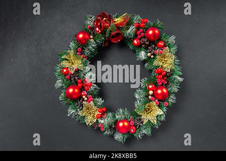 Una corona di Natale con un arco rosso e decorazioni di coni, agrifoglio, mistletoe, e verde invernale contro una porta di fronte di una quercia blu scuro. Foto Stock