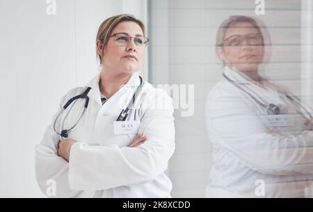 Riflettendo sui suoi giorni nella scuola medica... una giovane e attraente dottoressa che guarda premurosa mentre sta in piedi con le braccia incrociate nel suo ufficio. Foto Stock