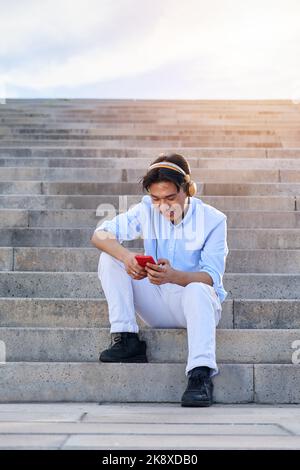 Allegro giovane asiatico che utilizza il telefono cellulare all'aperto, mentre si ascolta la musica, seduto su grandi scale. Foto Stock